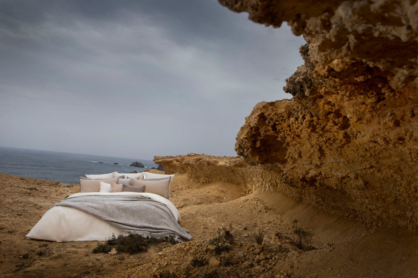 A bed with neutral-toned pillows is set up on a rocky coastline overlooking the ocean, featuring Panama Pillow Cover by Celso de Lemos available at Linen Society.