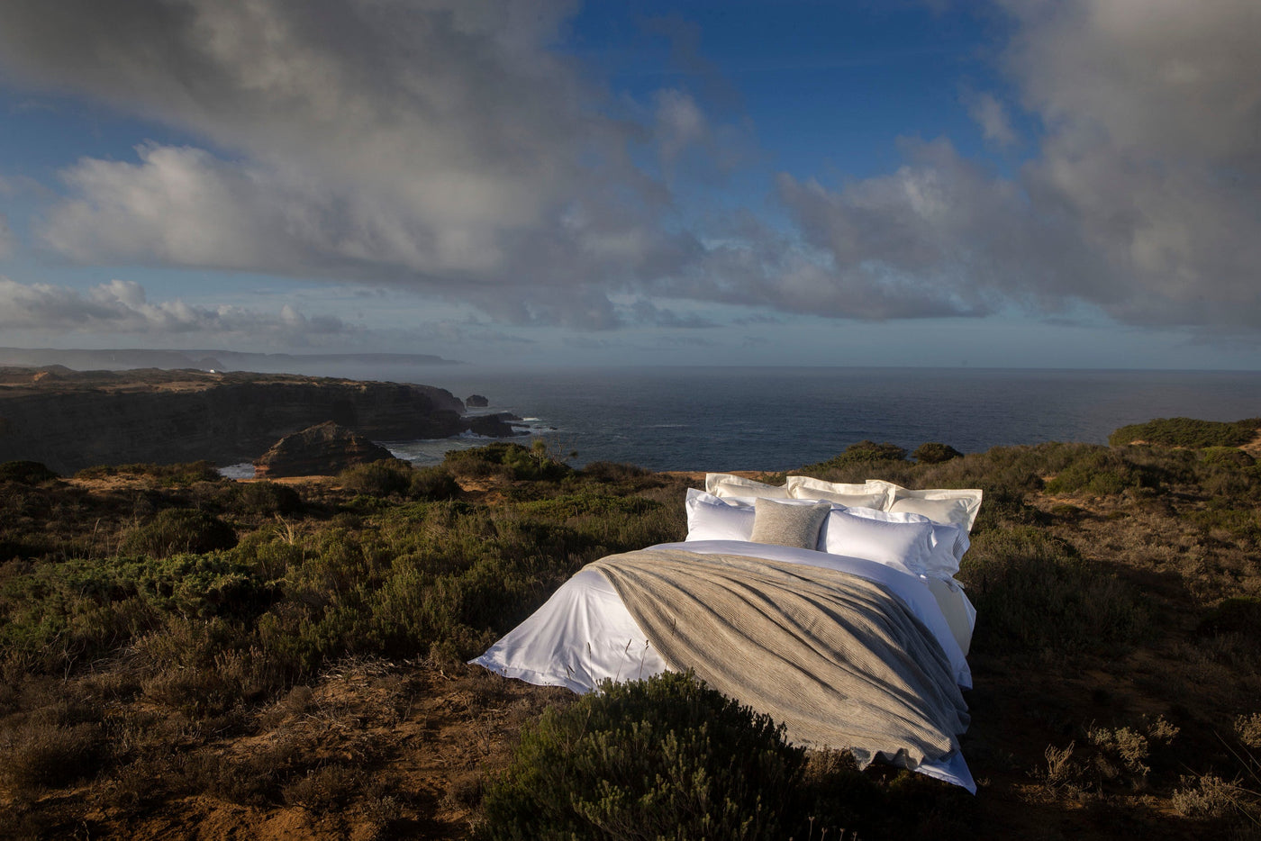 A neatly made bed with white linens and a textured throw rests on a coastal hillside overlooking the ocean, featuring Panama Pillow Cover by Celso de Lemos available at Linen Society.
