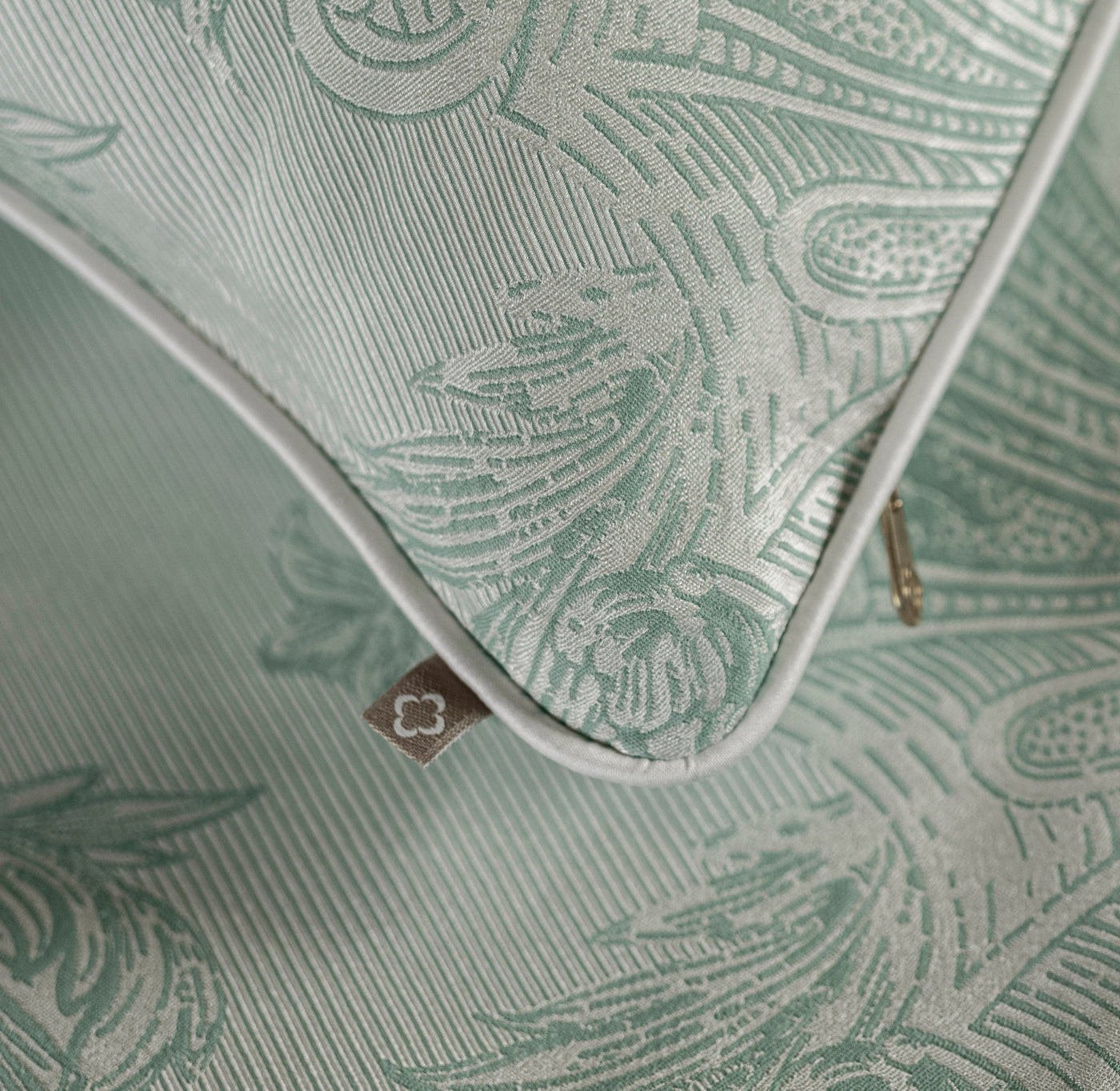 Close-up of sage green jacquard duvet cover corner, white piping and gold zipper detail visible.