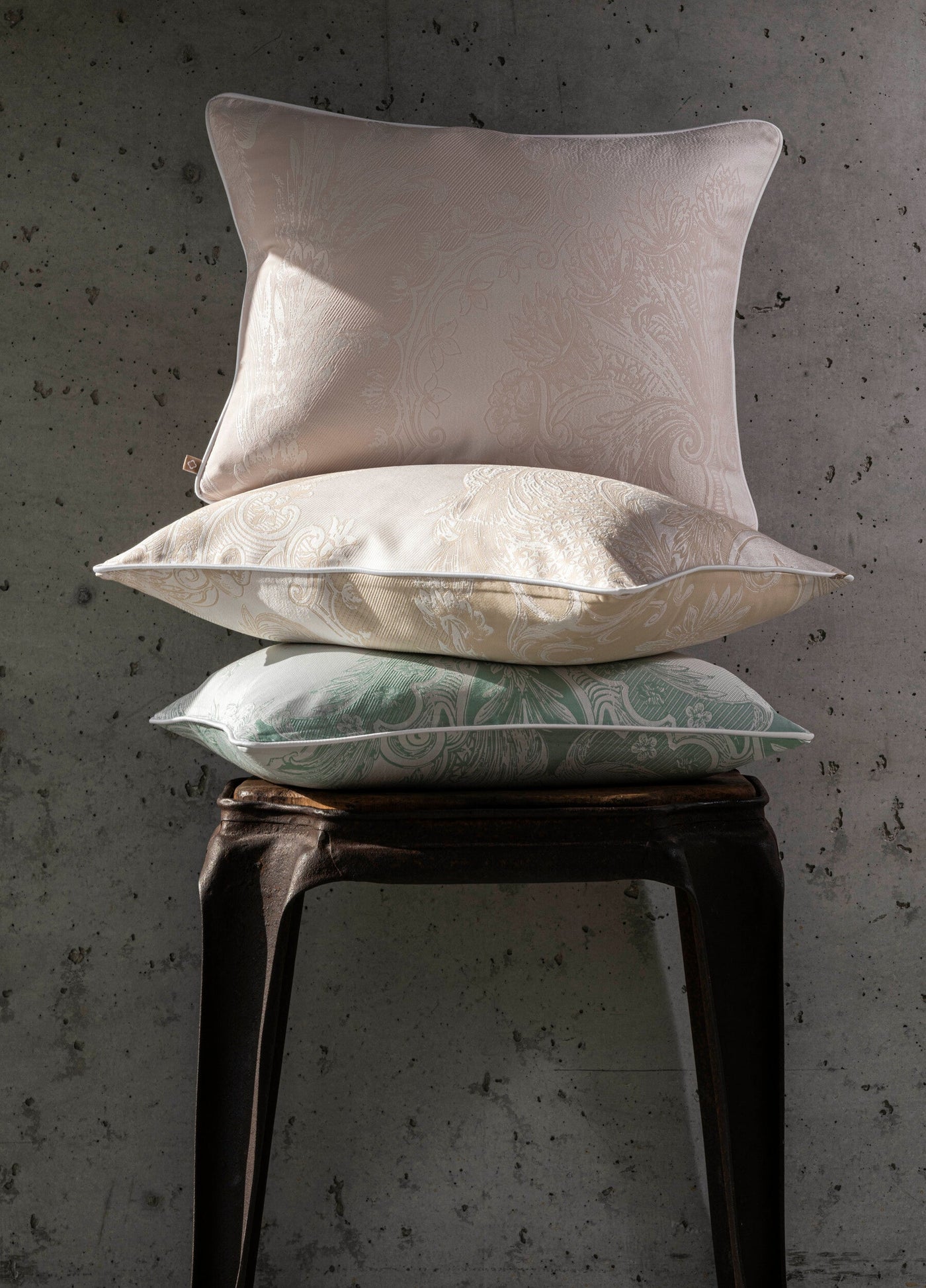 Three stacked Pompadour pillowcases in blush, ivory, and sage on a dark stool.