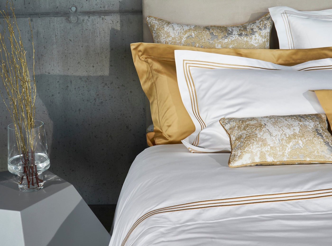 Styled bed with white flat sheet, gold embroidered border detail, against concrete wall.