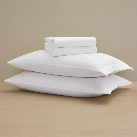 Two stacked white pillows and Frette’s Checkered Sateen Flat Sheet are arranged on a wooden surface, with the smooth bedding and sateen finish set against a beige backdrop.