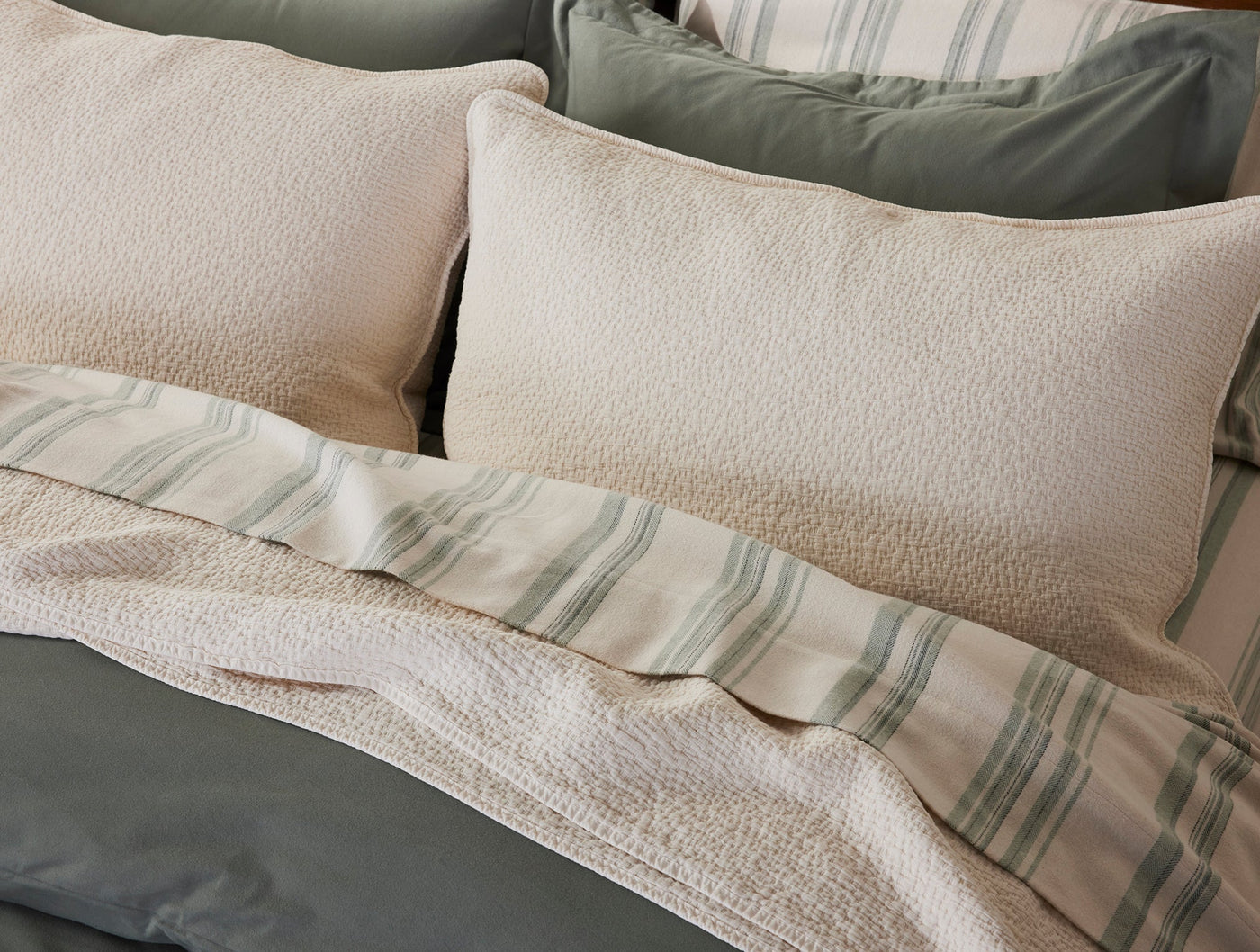 Ivory matelassé blanket draped over a bed with sage stripe and slate bedding layers.