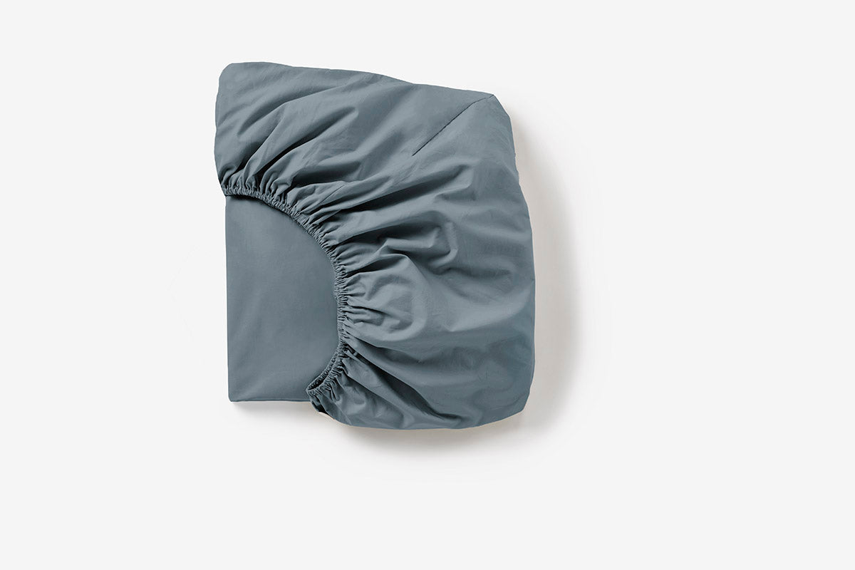 Coyuchi Cloud Soft Sateen fitted sheet folded, showing elasticated corners in slate blue.