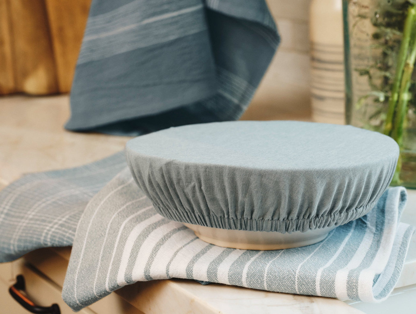 Coyuchi Conserve bowl cover in slate blue stretched over ceramic bowl, striped linen towels beneath.