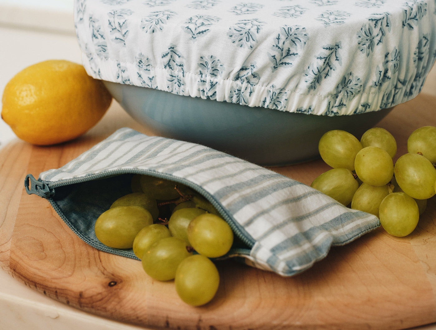 Striped Coyuchi snack bag filled with green grapes beside a floral bowl cover on a wood board.
