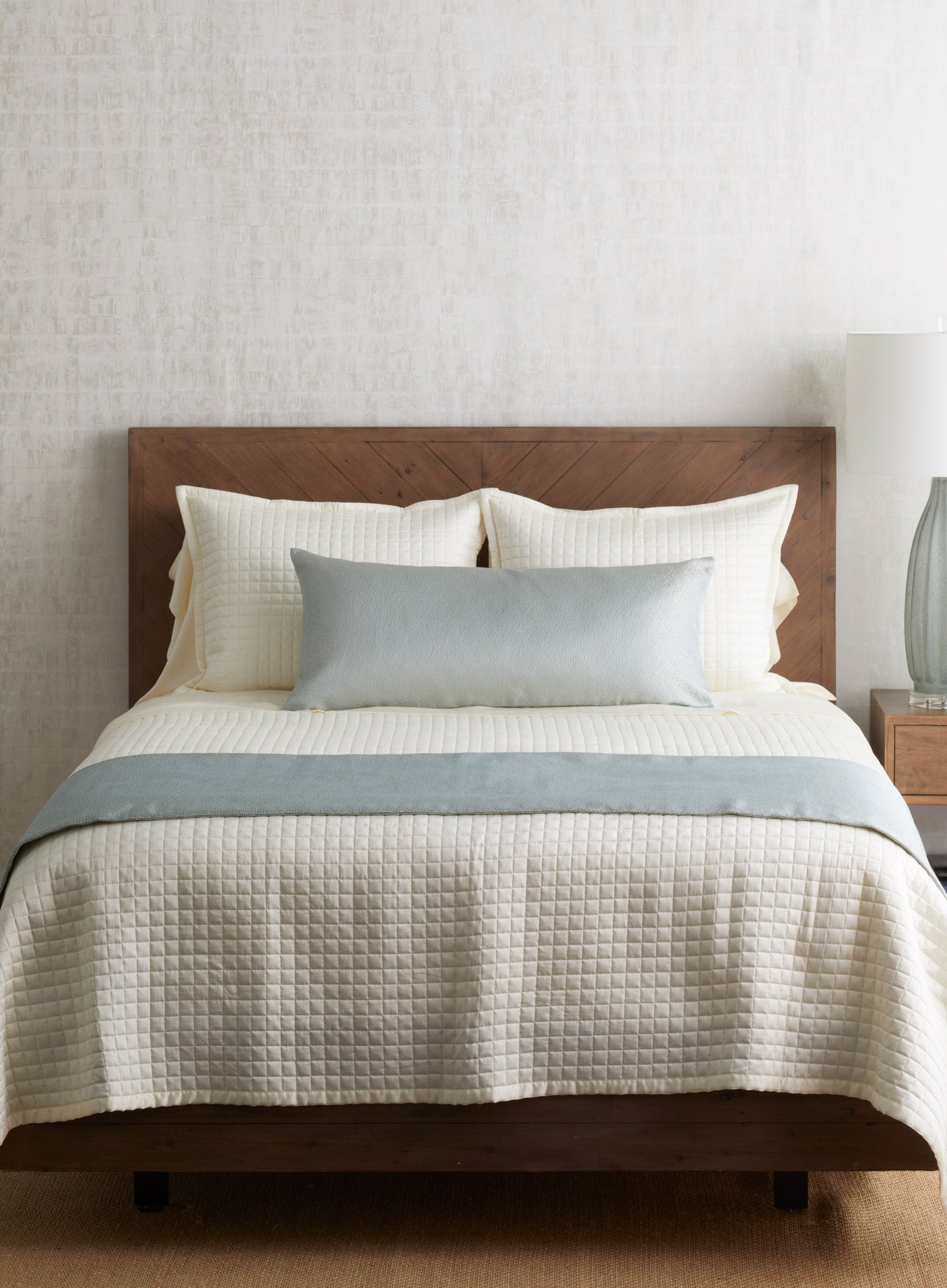 Ann Gish Dapple ivory quilted bed finisher styled on bed with dusty blue lumbar pillow accent.