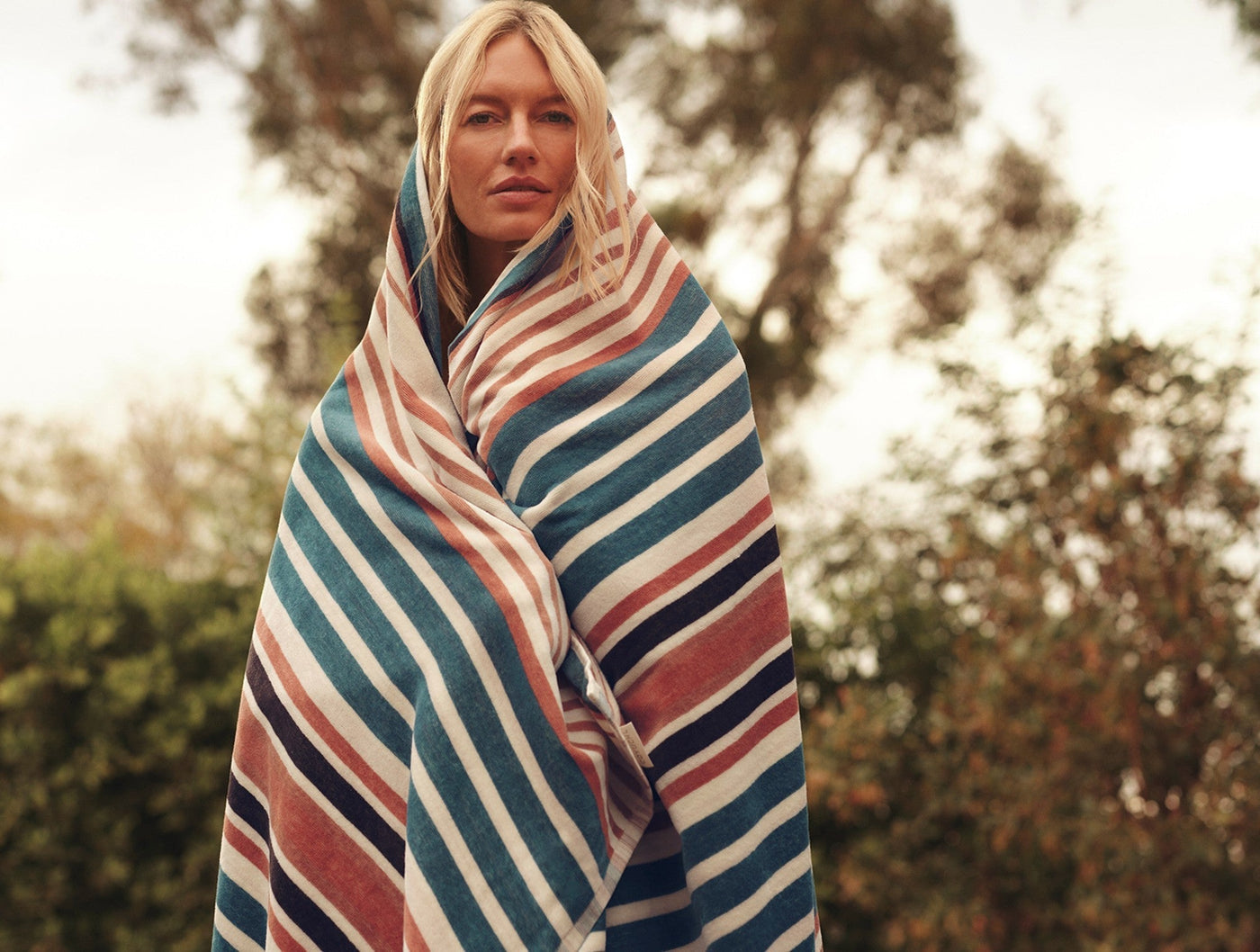 Woman wrapped in Coyuchi Morelia beach towel showing teal, terracotta, and ivory stripes outdoors.