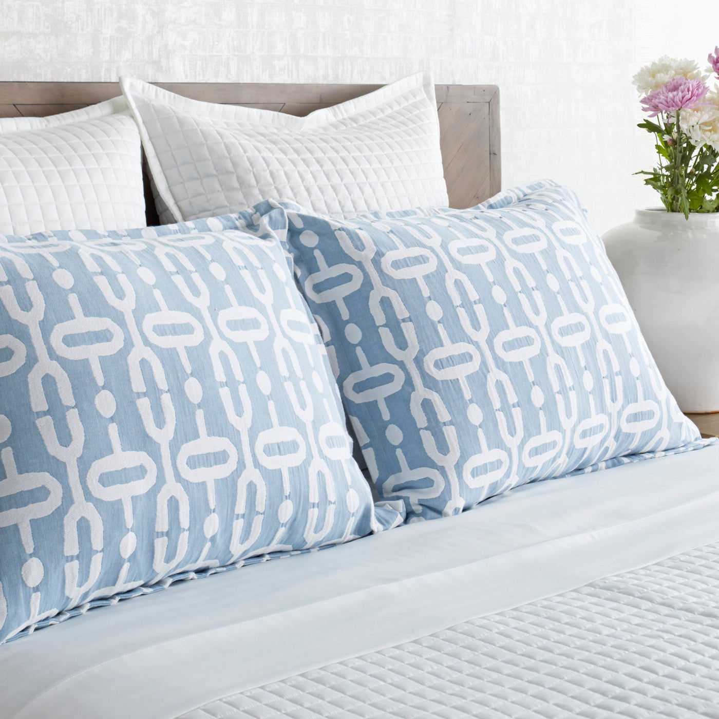 Two Ann Gish Doorknocker shams in sky blue with white geometric chain pattern on a styled bed.