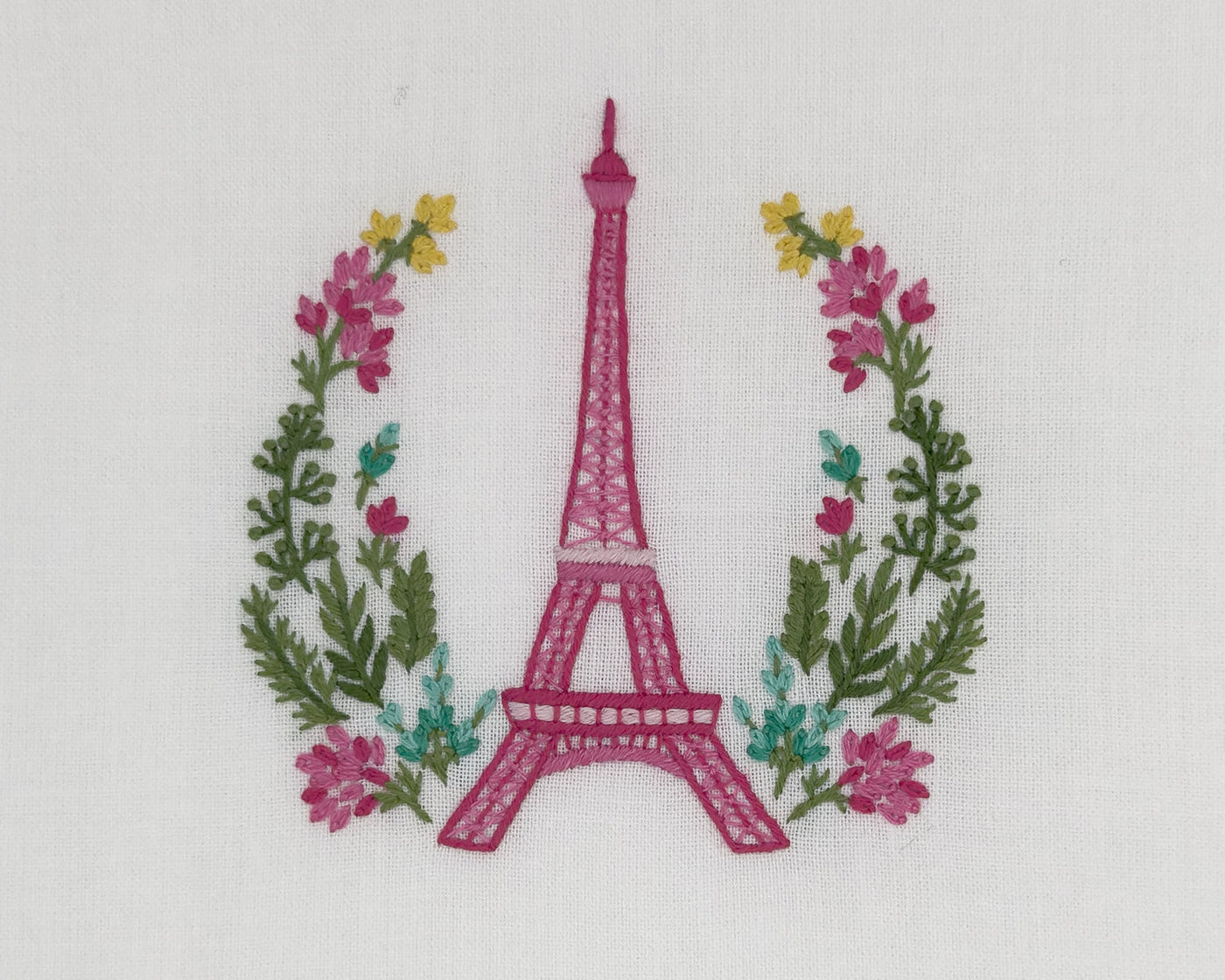Close-up of raspberry Eiffel Tower embroidery with floral wreath on white linen.