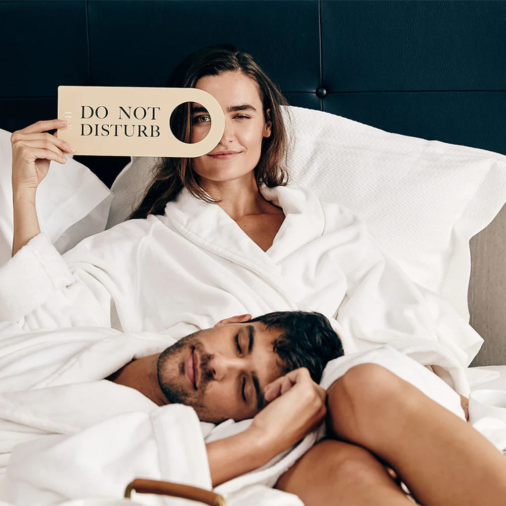 A woman in a white robe holds a Do Not Disturb sign, highlighting hotel luxury, while a man rests his head on her lap against a Frette Waves | Pique Euro Sham Set.