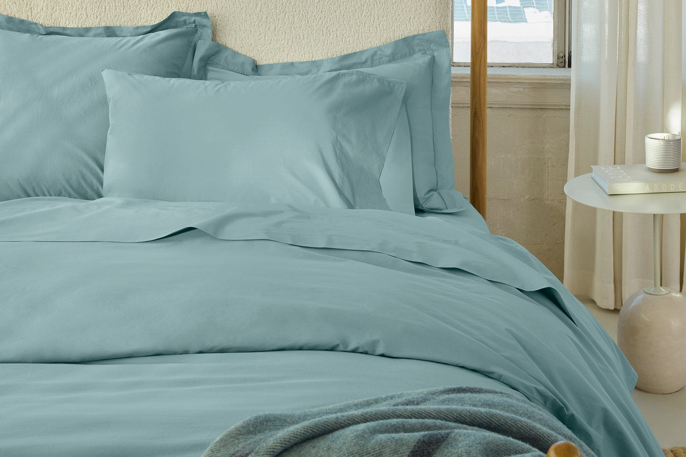 Close-up of Coyuchi Refined Percale sheet set in muted teal styled on a sunlit bed.