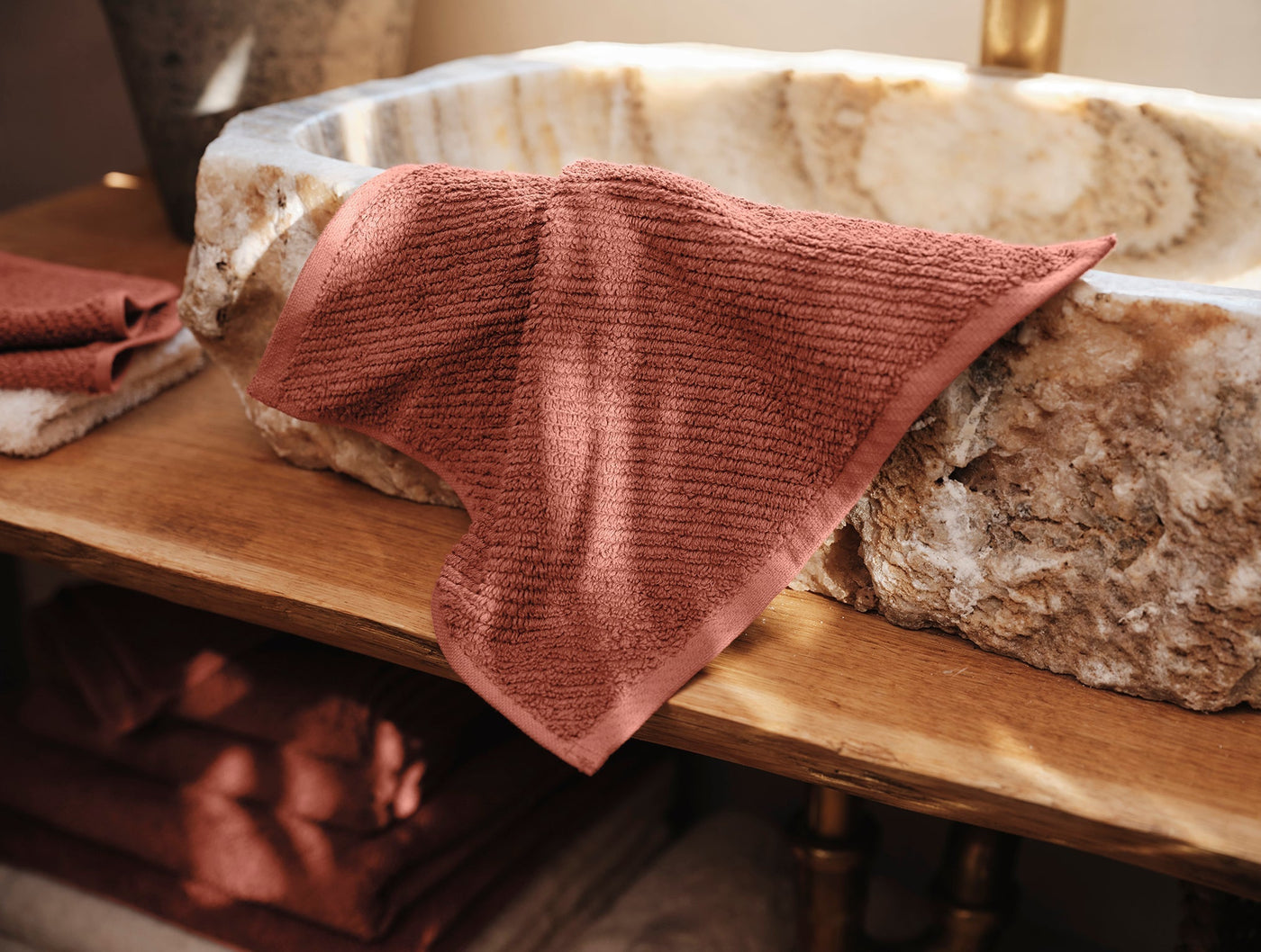 Coyuchi Air Weight wash cloth in terracotta, draped over a stone basin on a wood shelf.