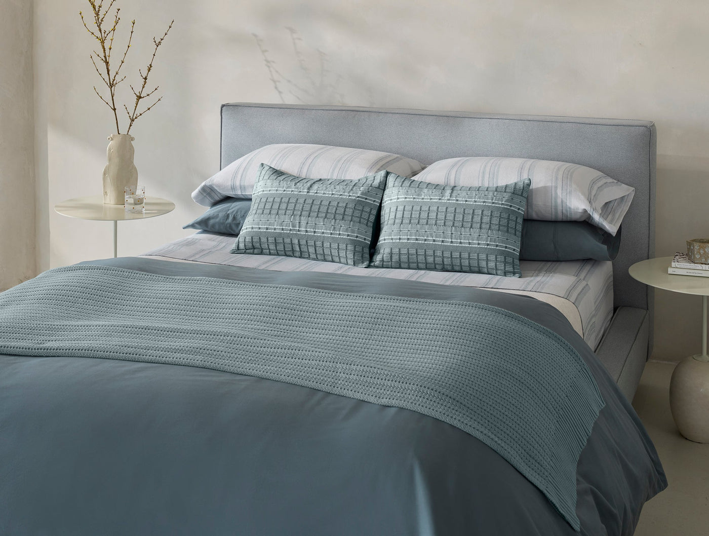 Slate blue knit throw draped over a made bed with layered bedding in a minimal bedroom.