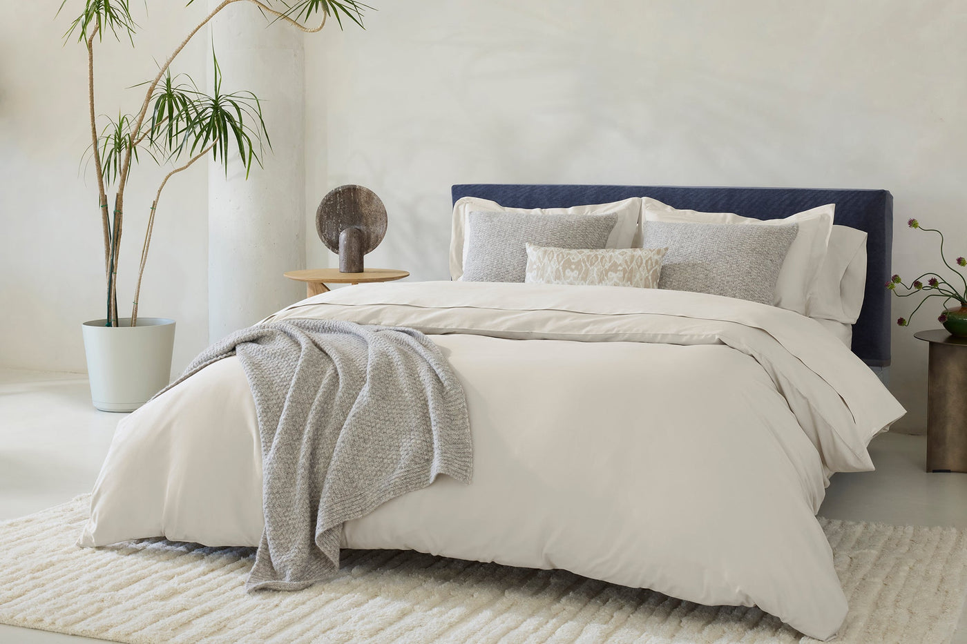 Ivory sateen sheet set styled on bed with navy headboard, gray throw, and patterned accent pillows.
