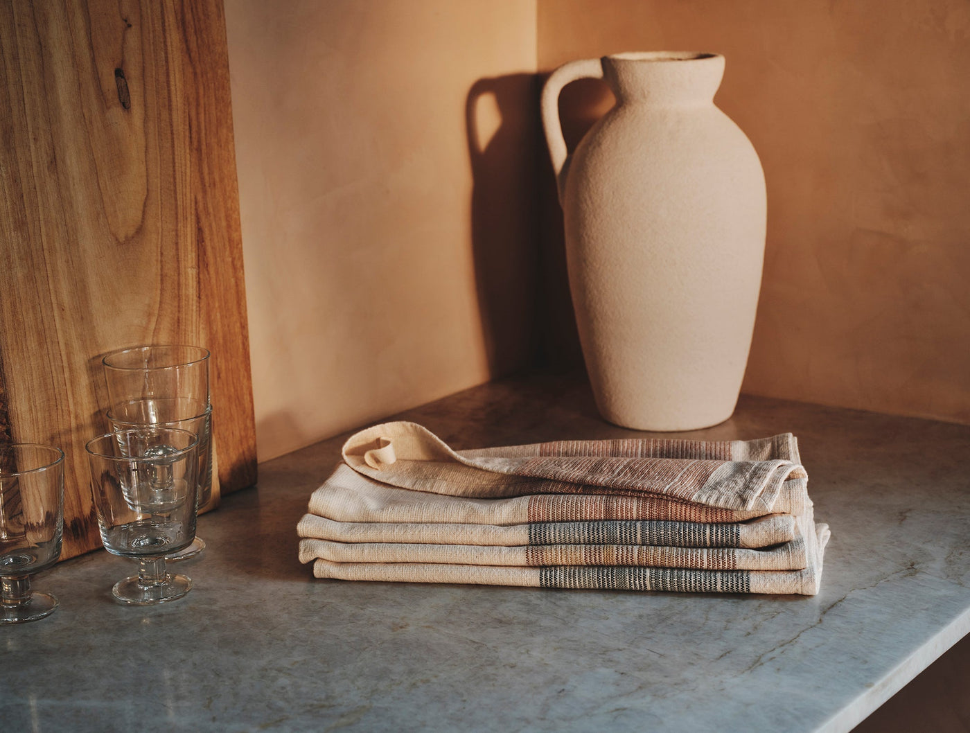 Folded Coyuchi Miraloma kitchen towels stacked on stone surface with ceramic jug and glassware.