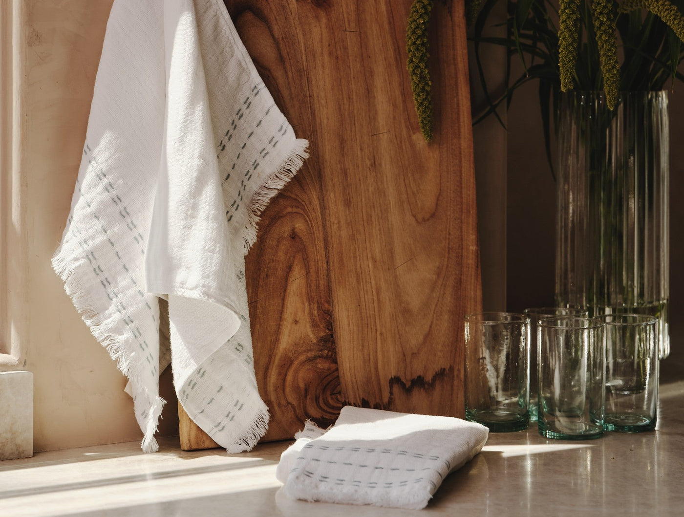 Coyuchi Alder white hand towels with fringe trim, draped and folded beside wood board and glassware.