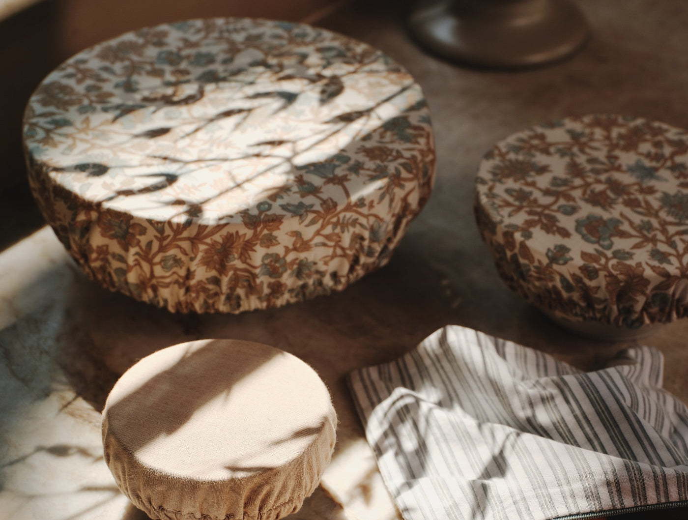 Three Coyuchi Conserve bowl covers in floral print, styled on a wooden tray with a striped linen cloth.