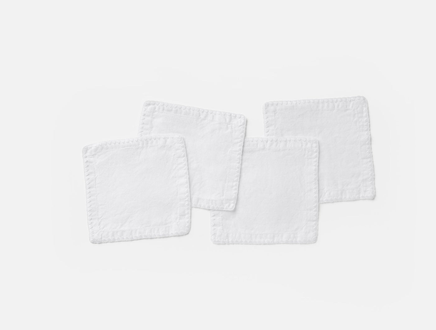 Four white Coyuchi Olivos linen cocktail napkins with embroidered trim, fanned on light background.
