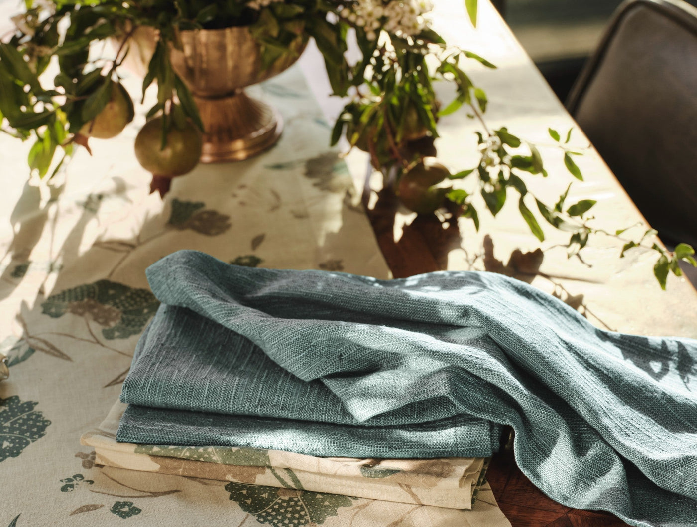 Folded slate blue textured linen napkins stacked on a floral tablecloth near a copper vase.