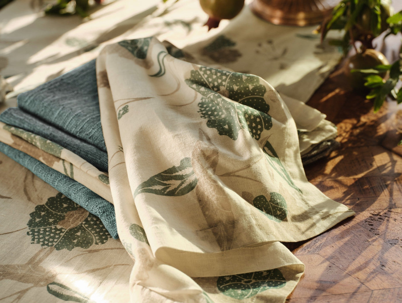 Ivory linen table runner with green botanical print, draped over stacked teal linens on a wooden surface.