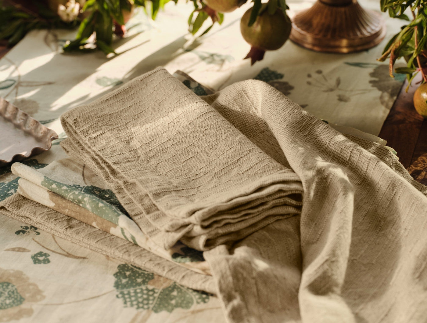 Folded natural linen napkins with fringe trim styled on a floral tablecloth, warm sunlight.