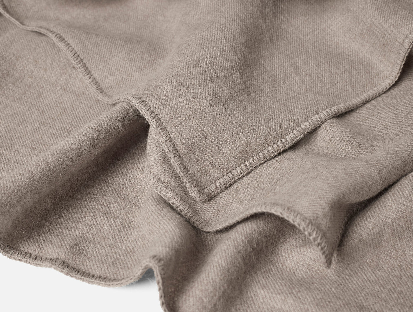 Close-up of taupe baby alpaca blanket corner showing overlock stitched edge and soft drape.