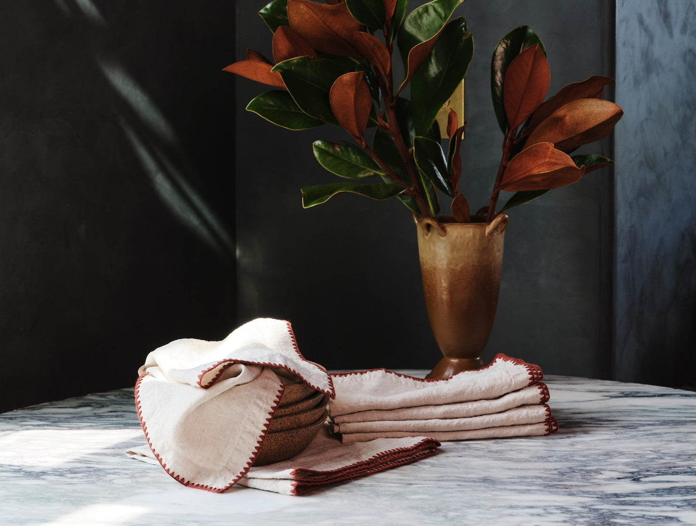 Ivory linen napkins with rust embroidered edges, folded on marble beside a brass vase.