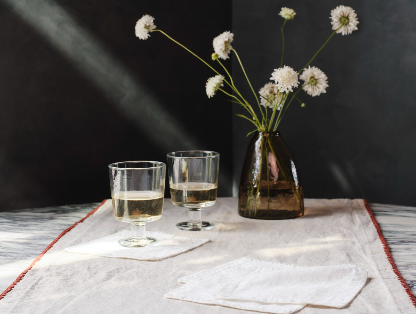 White linen cocktail napkins styled on marble with wine glasses and floral vase.