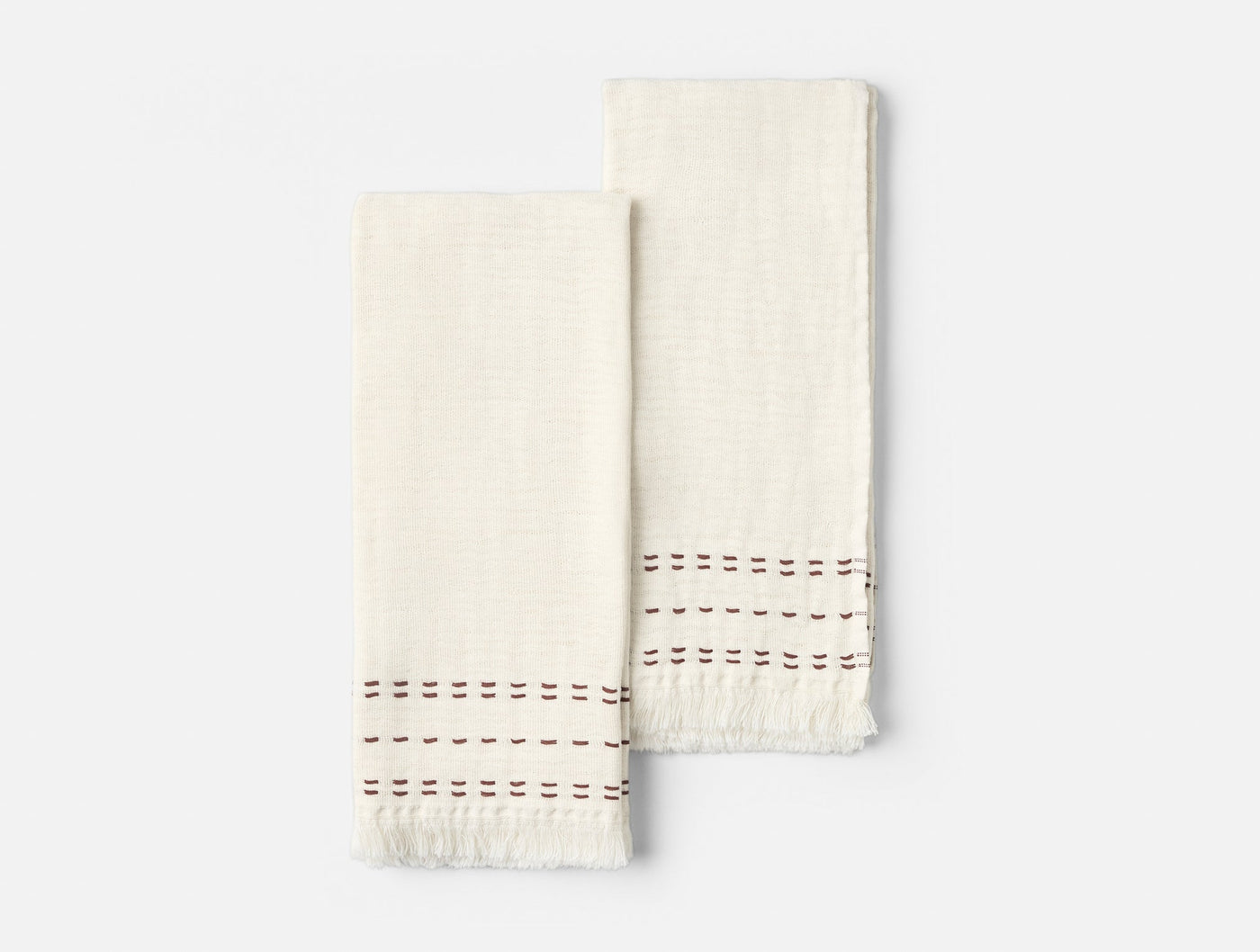 Pair of ivory hand towels with fringed hems and burgundy embroidered stitch border detail.