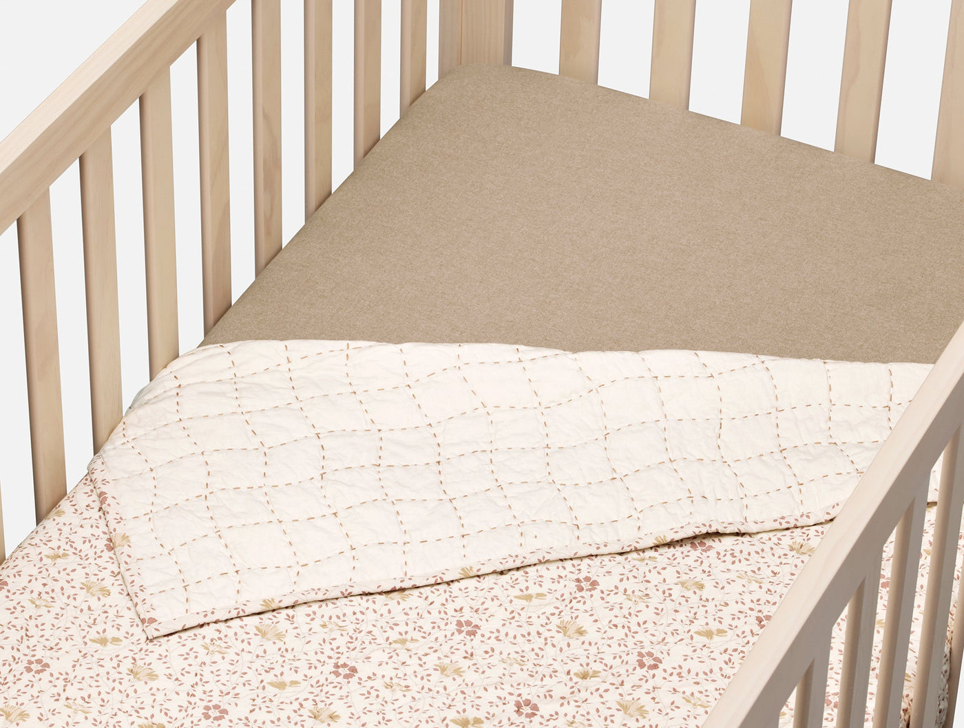 Coyuchi fitted sheet in tan flannel on a crib, with floral quilt folded at foot.
