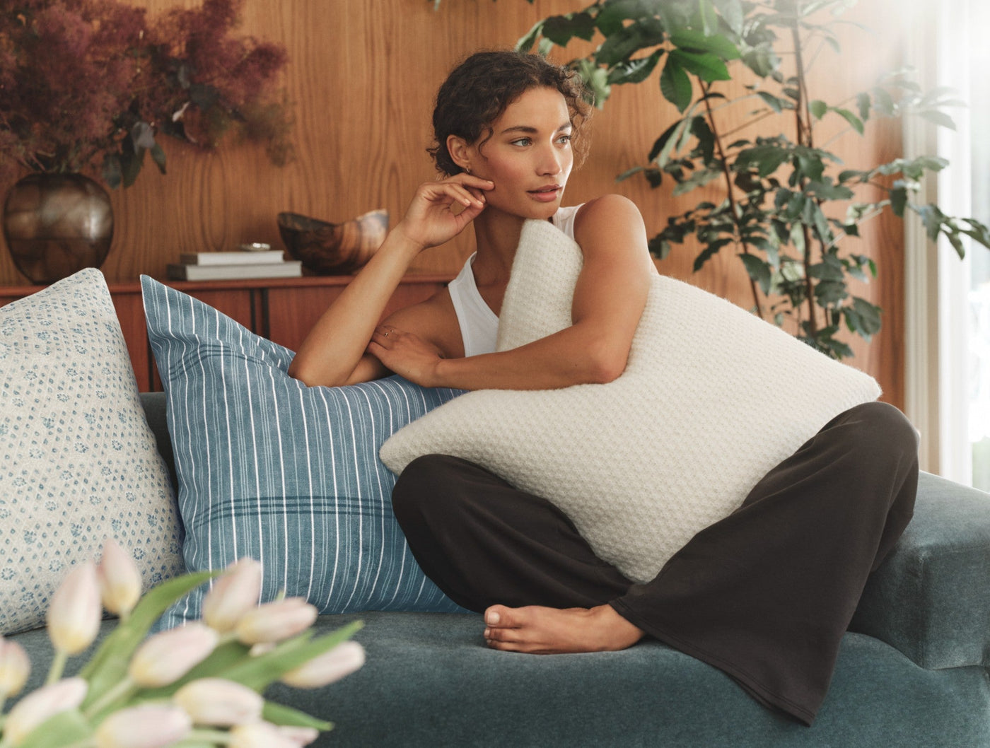 Woman seated on teal sofa hugging ivory textured Coyuchi Fernwood pillow, blue plaid pillow beside her.