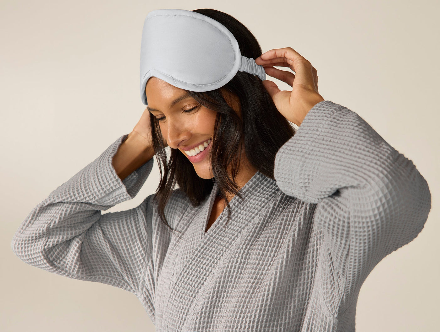 Smiling woman adjusting a Coyuchi silver grey eye mask, wearing a grey waffle robe.