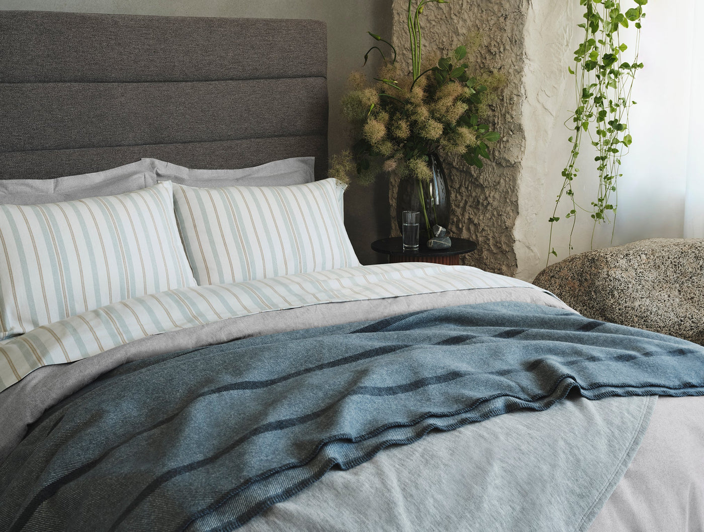 Denim blue brushed flannel throw draped over a made bed with striped pillowcases, rustic stone wall backdrop.