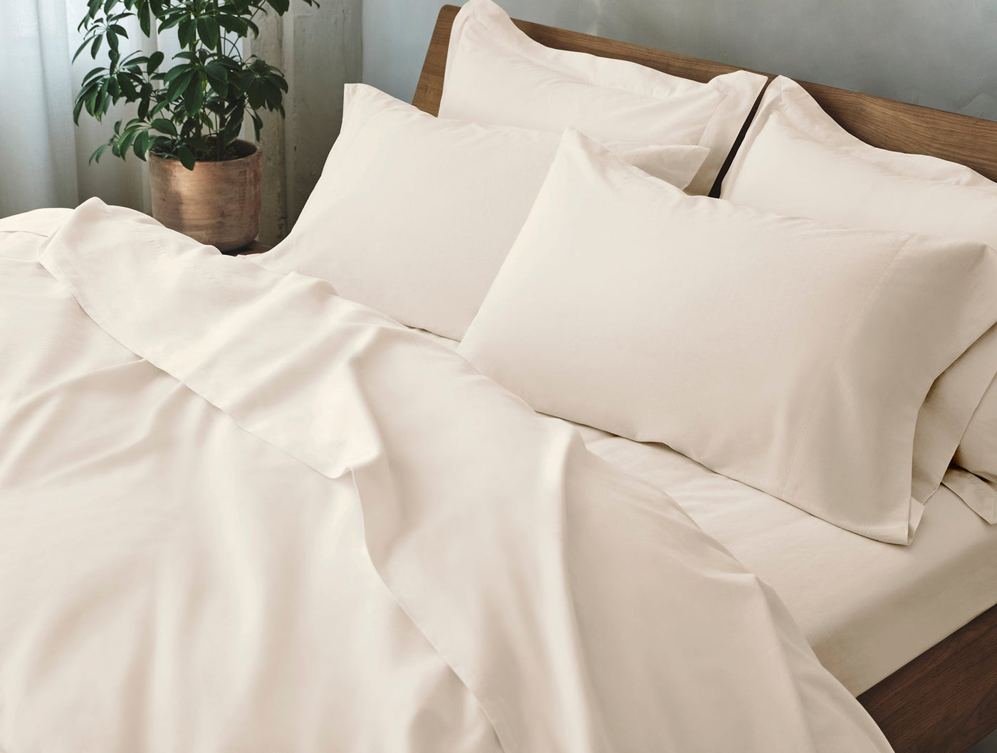 Ivory sateen sheet set draped on wood-framed bed with layered pillows and plant.