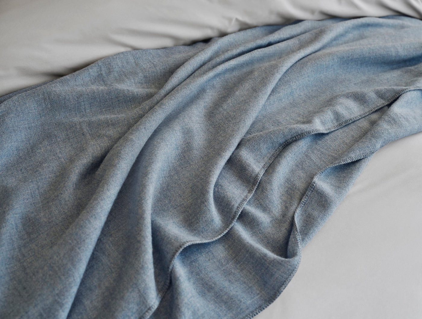 Slate blue alpaca throw draped loosely across white bedding, showing soft drape and woven texture.