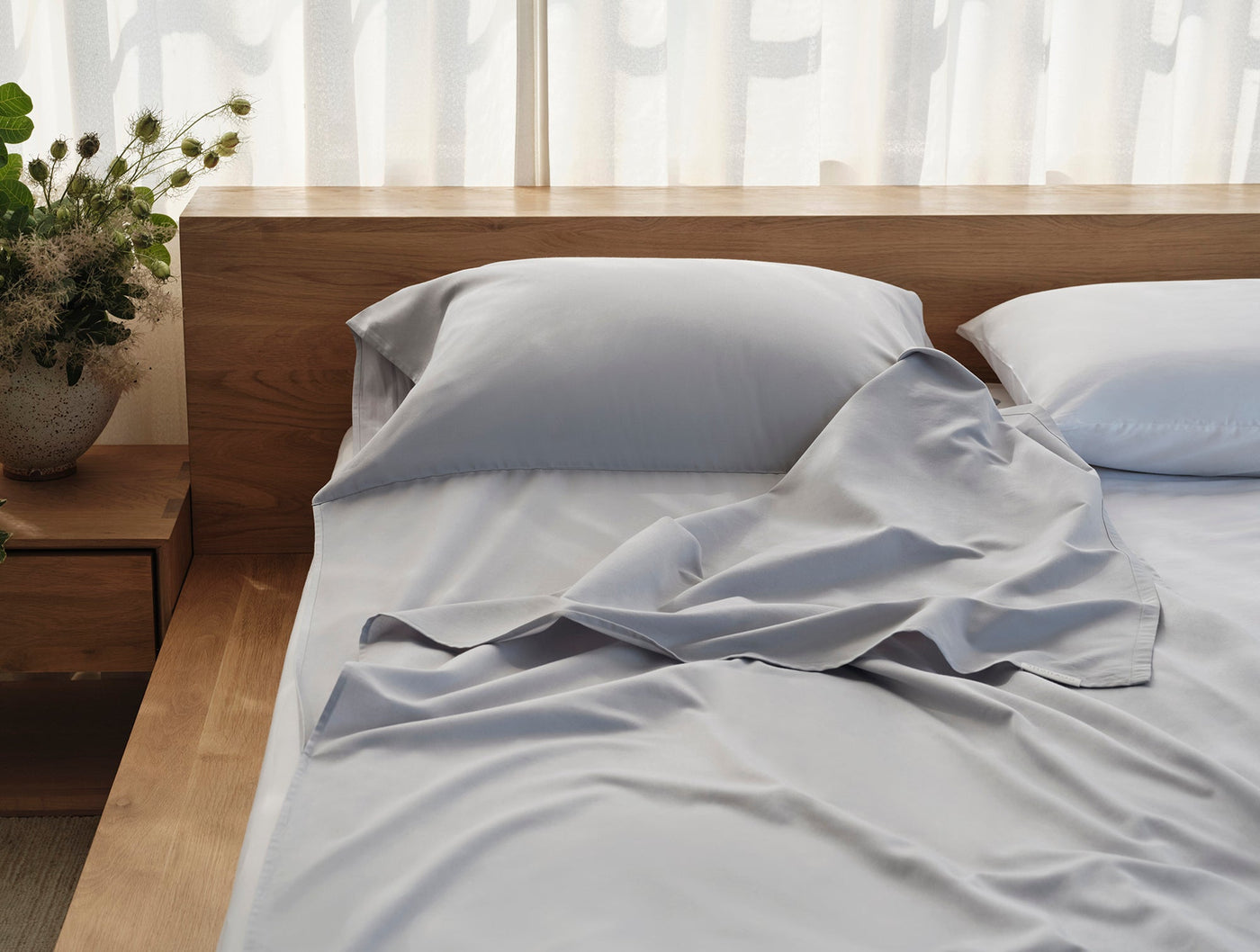 Loosely draped silver-blue sateen sheets styled on a wood-framed bed in a sunlit room.