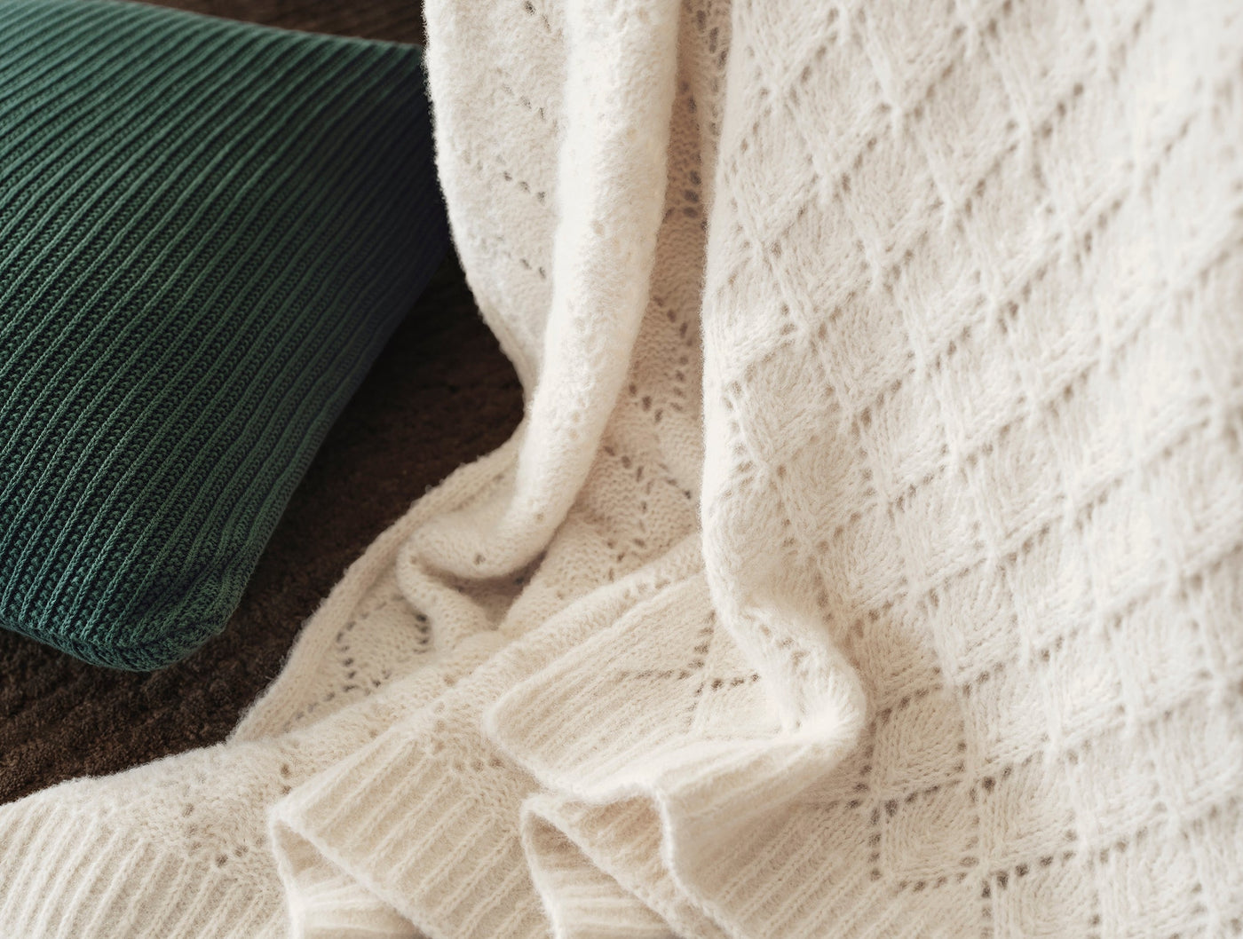 Close-up of ivory alpaca cotton knit throw with open diamond pattern, green ribbed pillow behind.