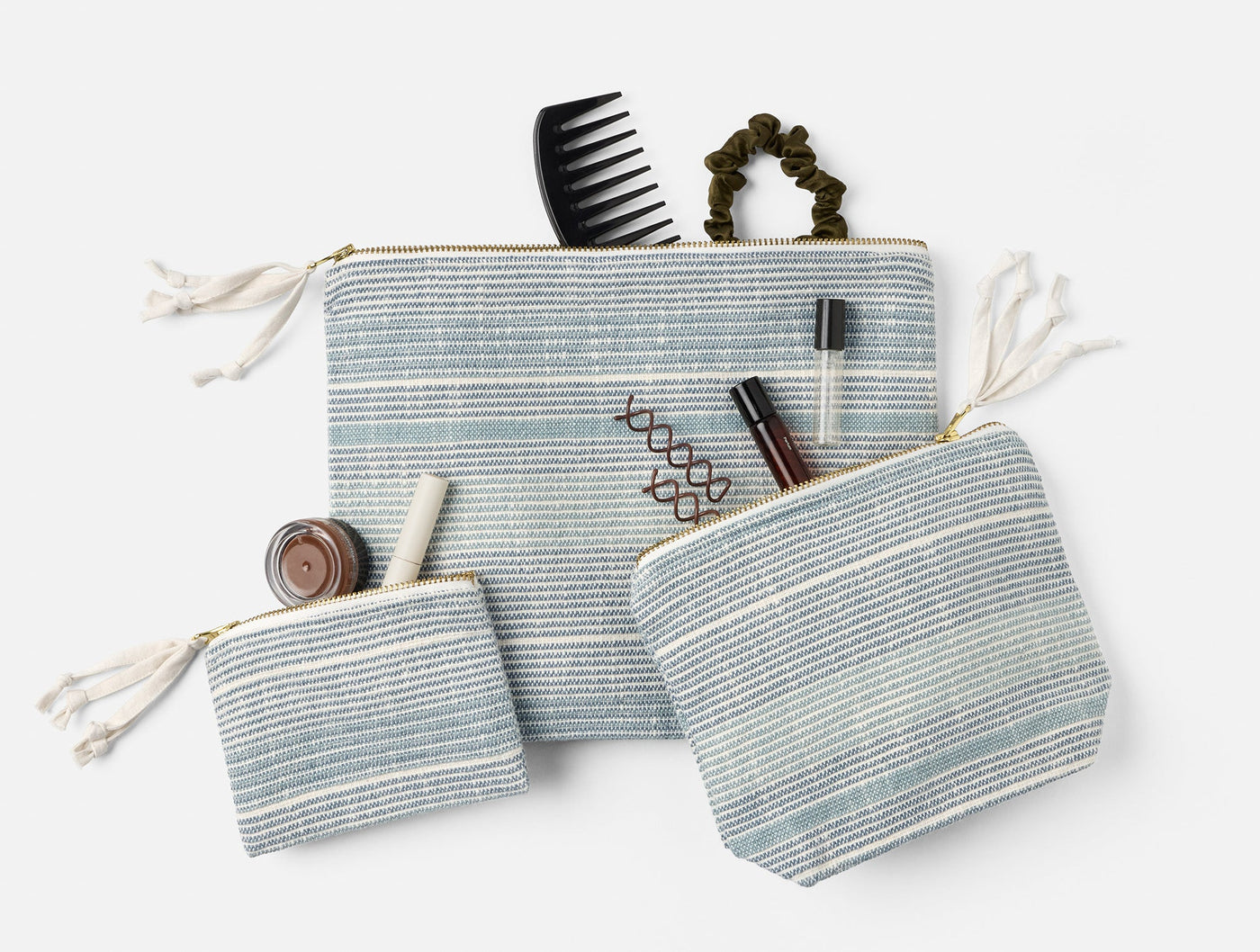 Three Coyuchi Miraloma pouches in muted blue stripe, styled open with beauty accessories.