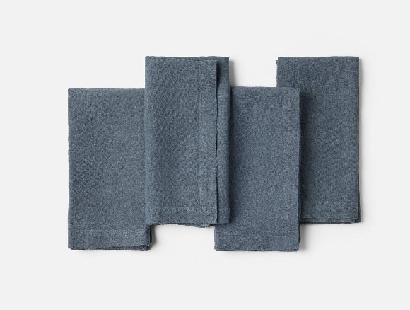 Four slate blue linen napkins arranged in a flat lay, slightly overlapping on a white background.