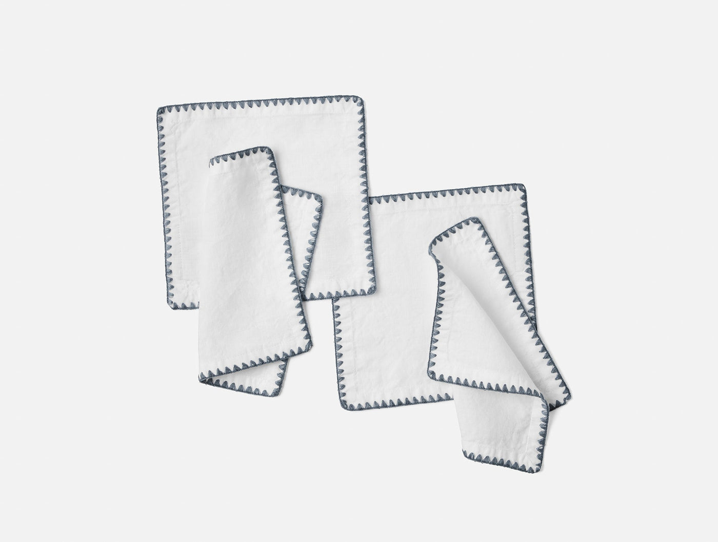 Coyuchi Olivos cocktail napkins fanned flat, white linen with slate embroidered edge trim.