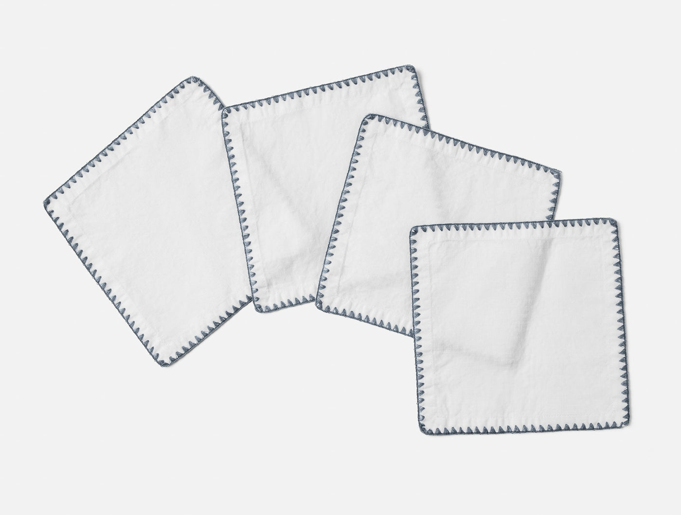 Four white Coyuchi Olivos linen napkins with slate blue embroidered border trim, fanned flat.