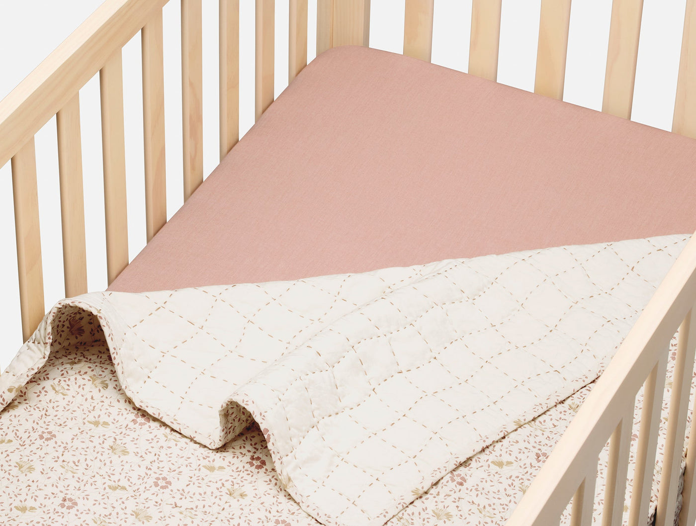 Dusty rose fitted sheet on crib mattress with quilted ivory blanket folded back.