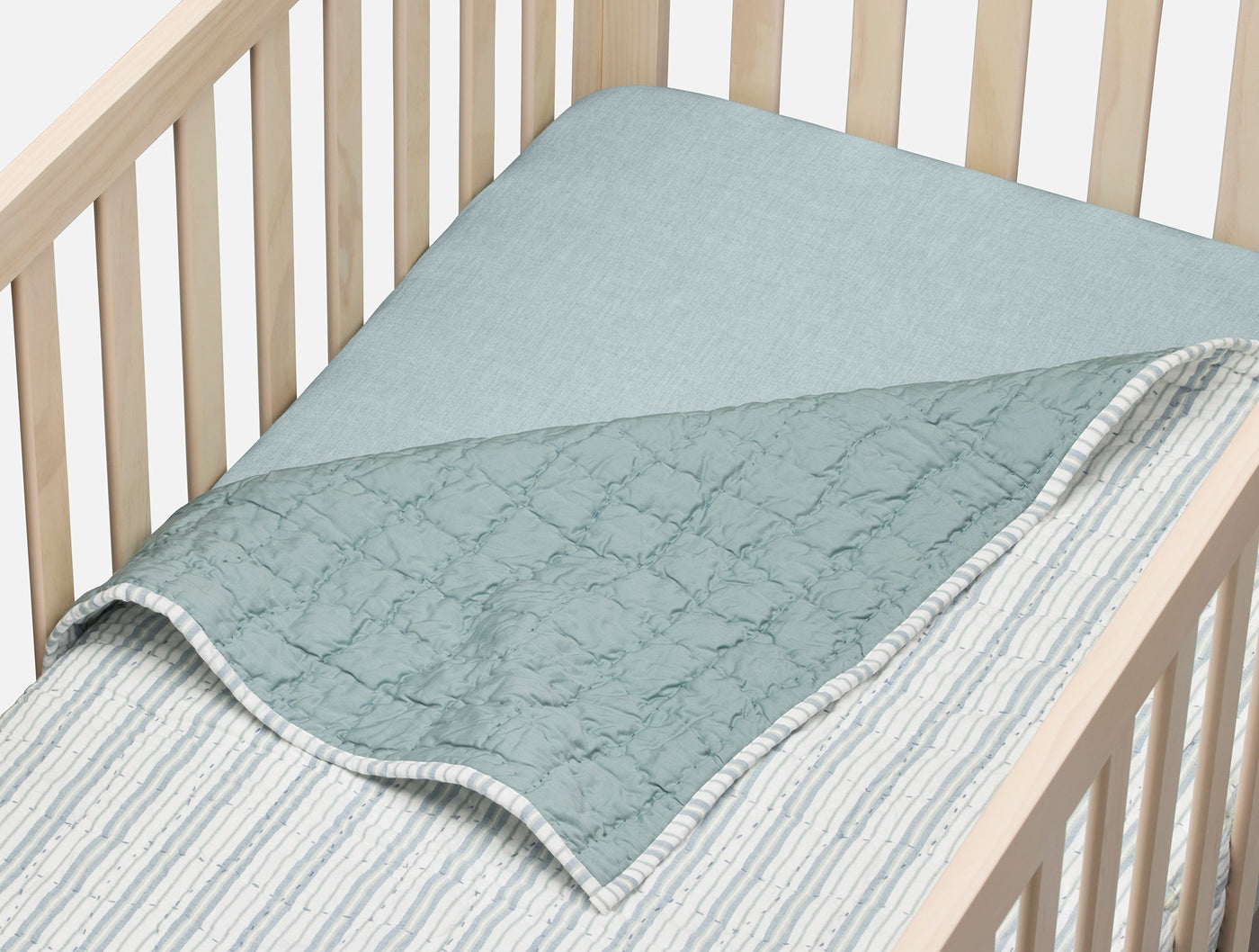 Sage green quilted blanket folded back in a natural wood crib, revealing striped percale lining.