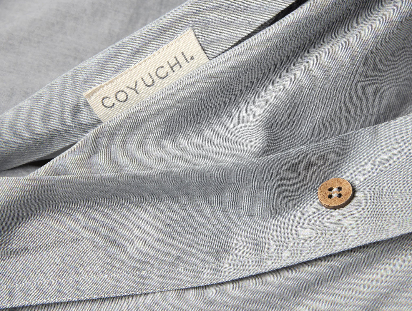 Close-up of Coyuchi duvet cover hem with wooden button closure and woven label, slate blue.