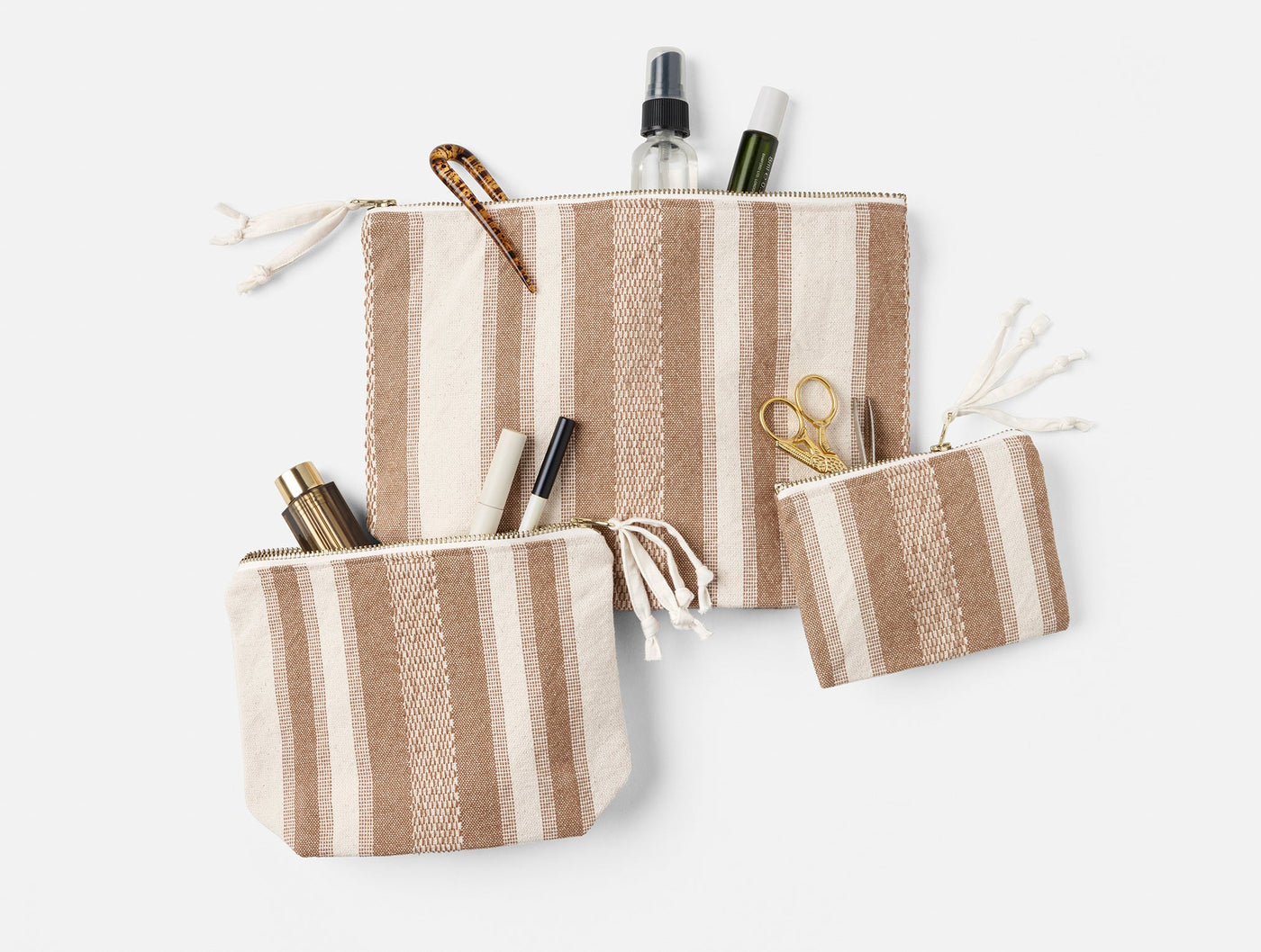 Three Coyuchi Serrano pouches in tan stripe, shown open with beauty accessories inside.