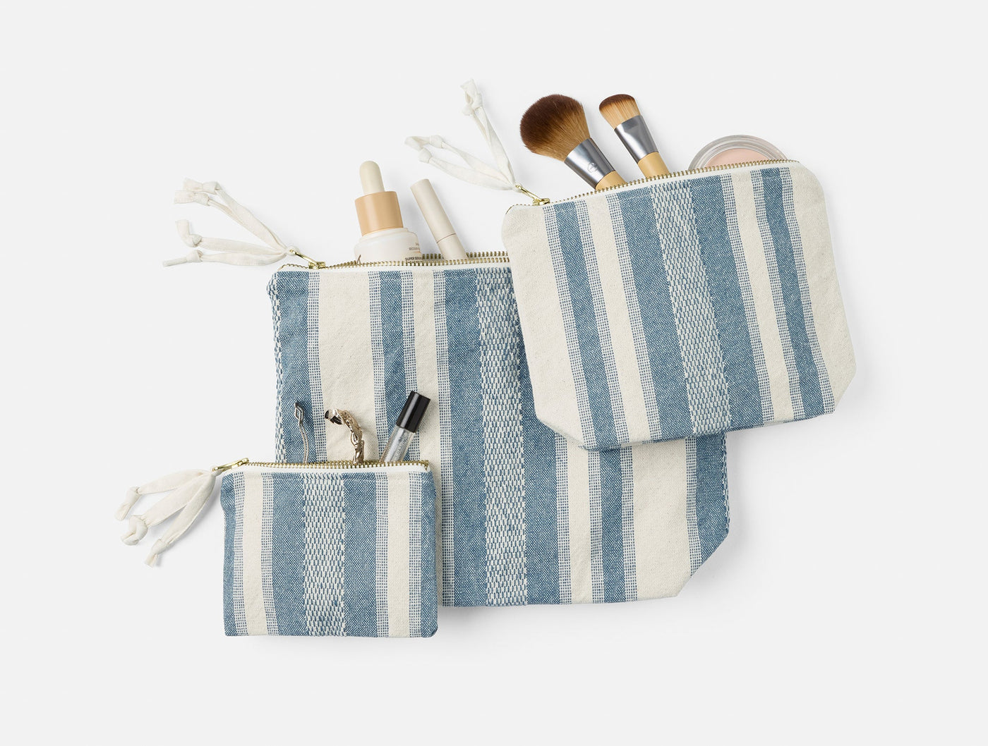 Three Coyuchi Serrano pouches in dusty blue stripe, styled open with makeup brushes and beauty tools.