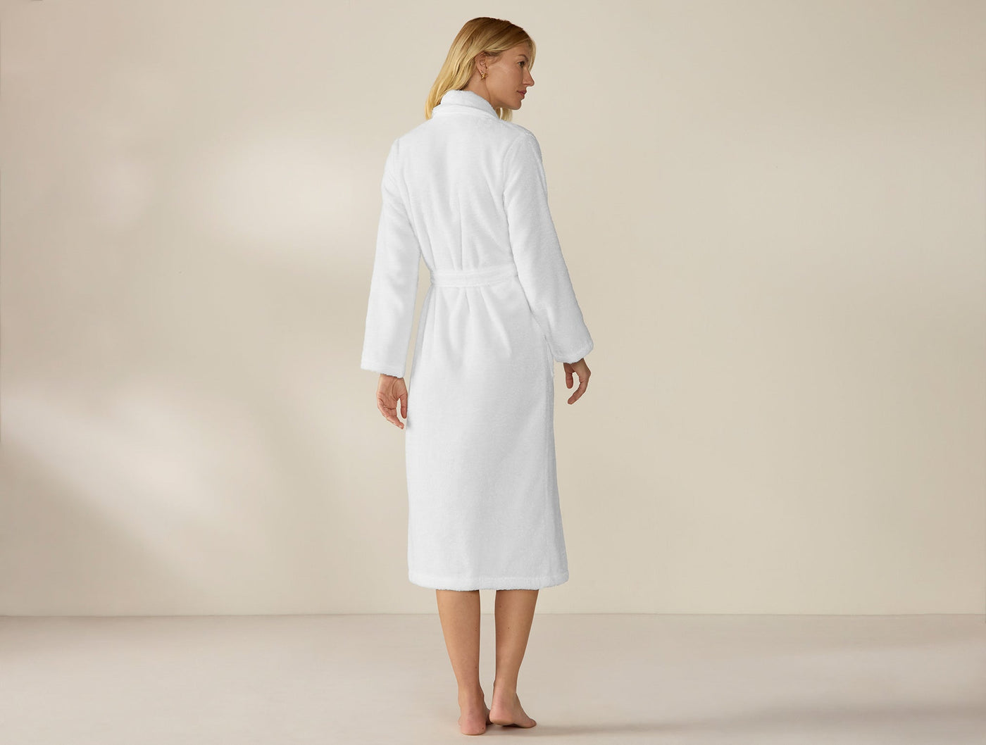 Rear view of white terry robe with belted waist, mid-calf length, worn by standing model.