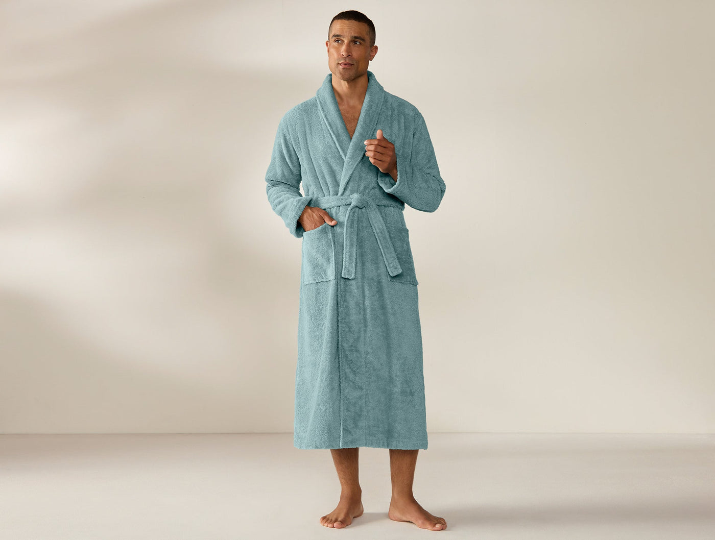 Man wearing full-length Coyuchi Cloud Loom robe in sea blue terry, belted, shawl collar.