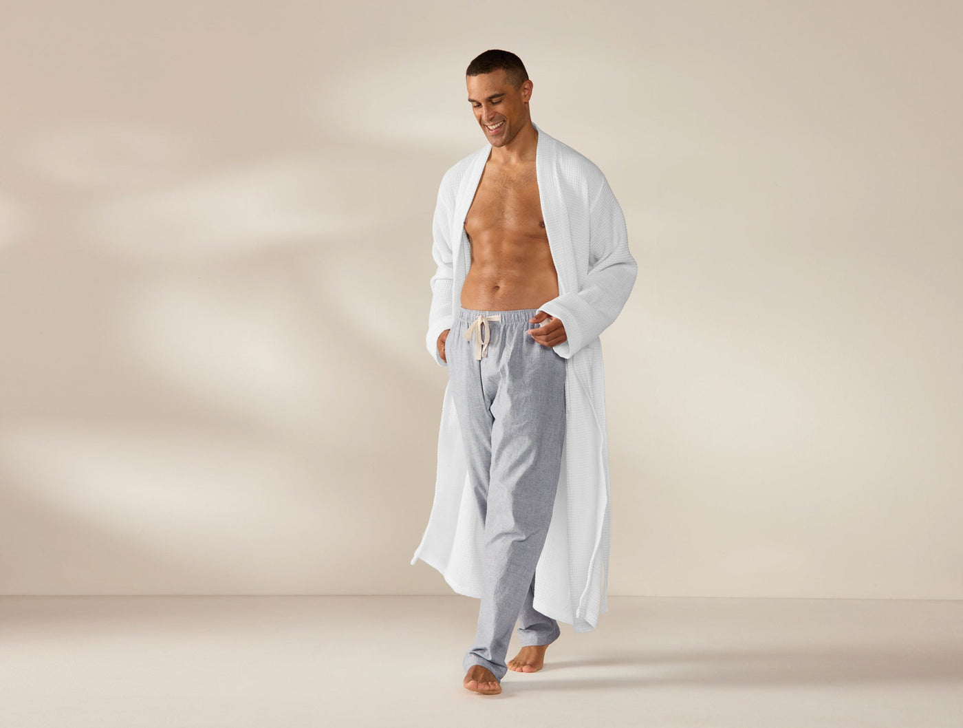 Man wearing open Coyuchi white waffle robe with grey drawstring pants, walking against warm neutral backdrop.